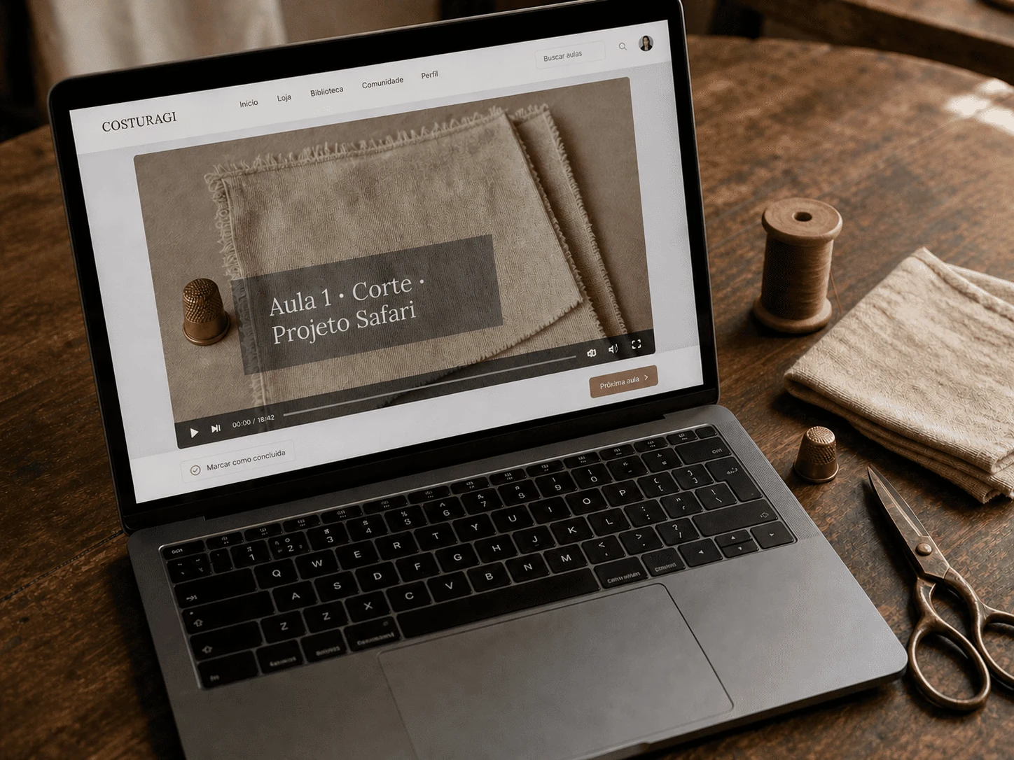 MacBook em bancada de ateliê mostrando aula do Projeto Safari · interface Hotmart com vídeo da Gi