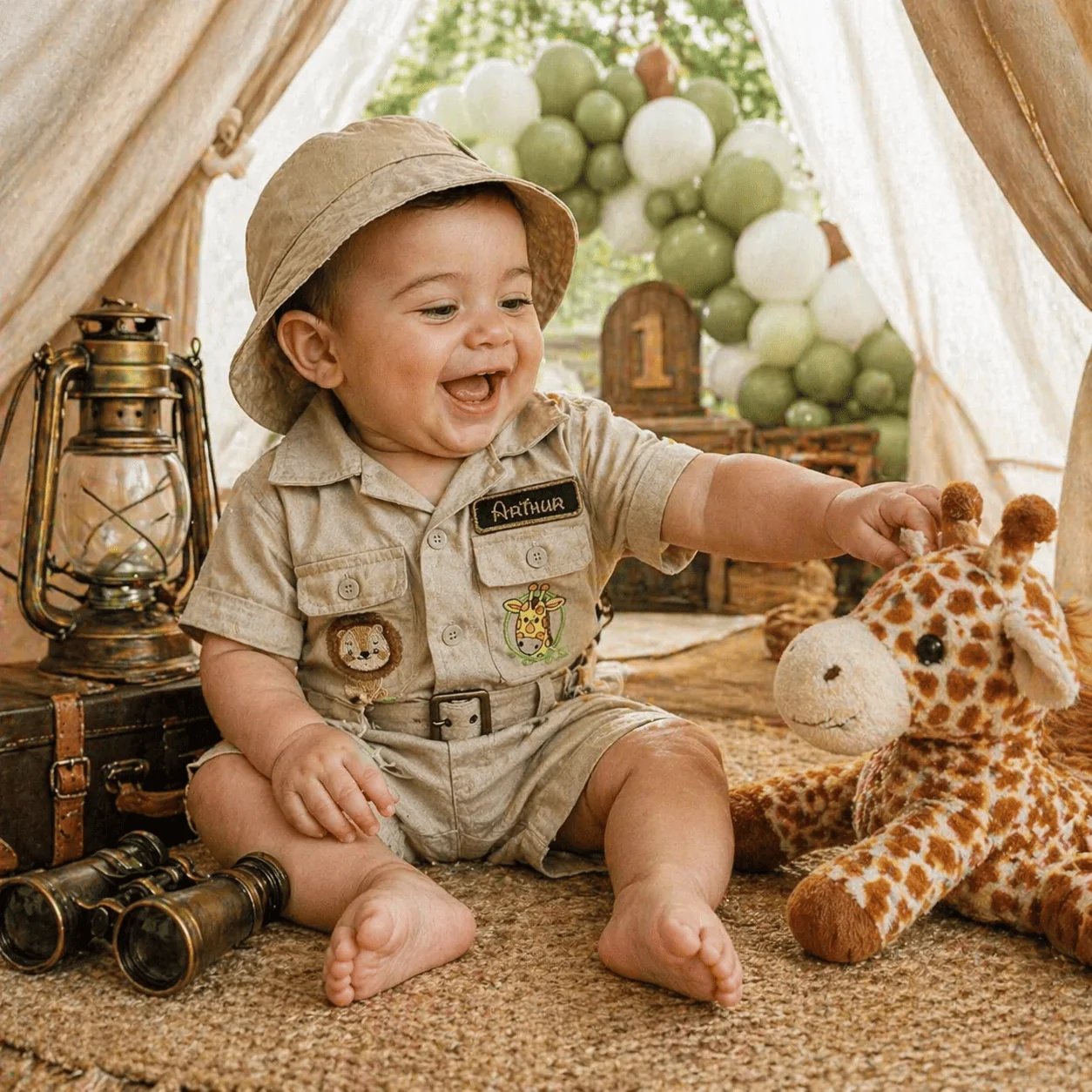 Bebê Arthur (~1 ano) gargalhando em ensaio fotográfico temático dentro de tenda de lona bege — vestindo Macacão Safari com etiqueta Arthur bordada + chapéu boonie · girafa de pelúcia ao lado, lanterna a óleo, binóculos vintage, banner com número '1', arco de balões verde oliva + marfim ao fundo · ambientação de mesversário temático