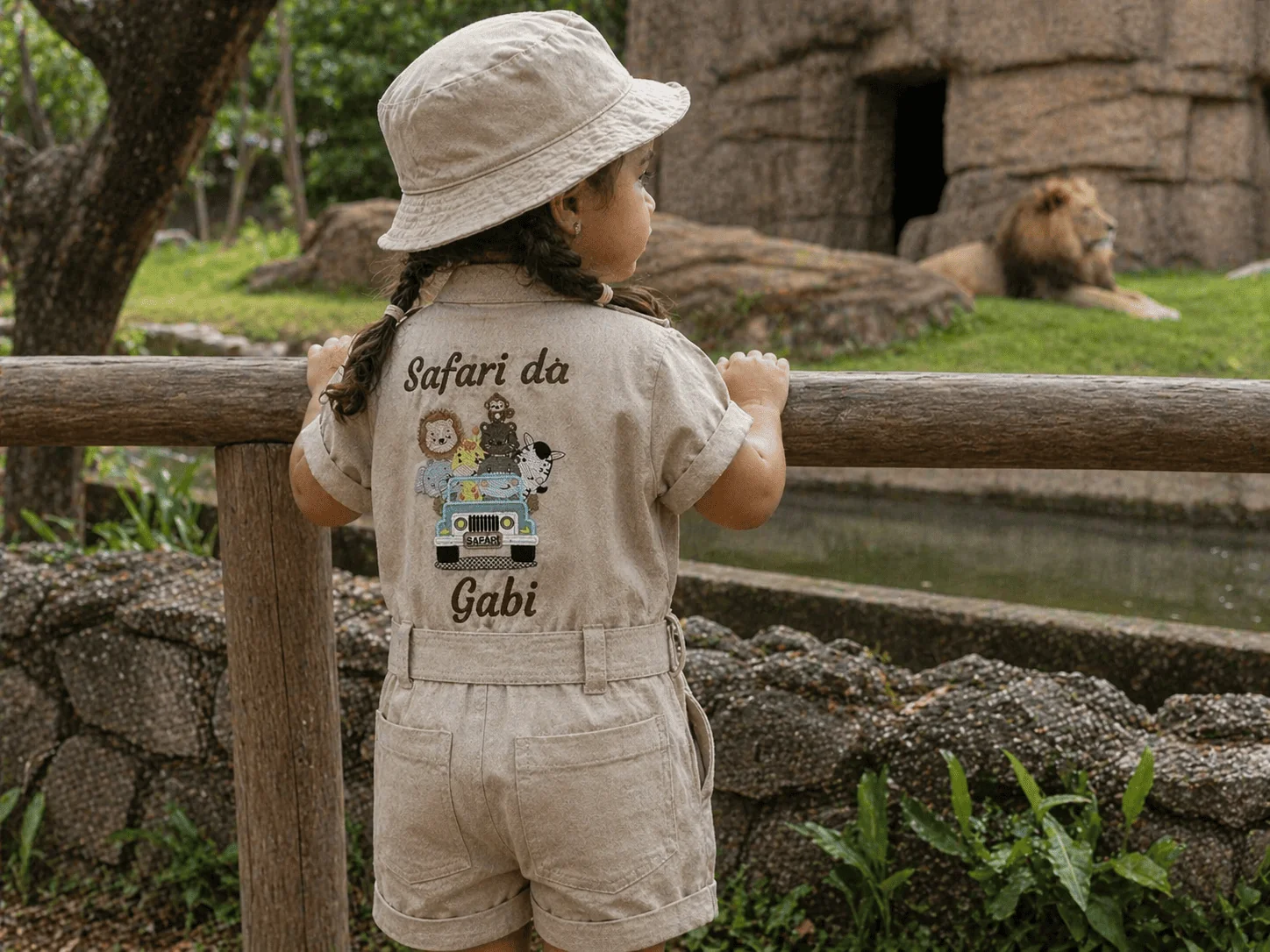Menina com chapéu boonie e trança vestindo Macacão Safari em linho cru com bordado 'Safari da Gabi' nas costas — encostada em grade de zoológico observando leão real ao fundo · personalização e tema confirmando a narrativa Safari
