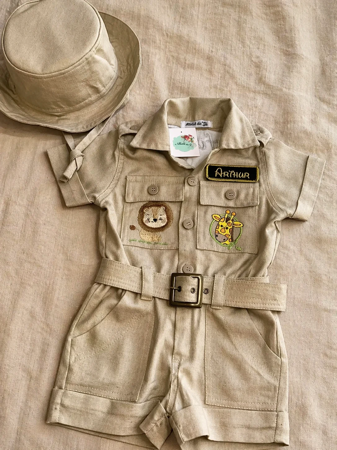 Flat-lay editorial do Macacão Safari na cor linho bege sobre fundo de linho cru · chapéu boonie combinando no canto superior esquerdo · etiqueta bordada 'Arthur' em caixa preta com borda oliva · bolso esquerdo com bordado de leão feito à mão · bolso direito com bordado de girafa em círculo verde · 5 botões de madrepérola bronze · cinto de linho com fivela quadrada bronze · dois bolsos frontais com pesponto duplo · luz warm natural lateral · fotografia editorial Kinfolk