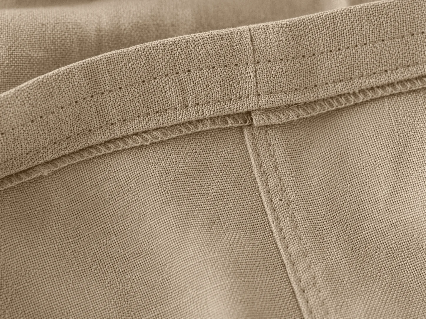 Macro extremo do avesso do Macacão Safari em linho bege natural mostrando o acabamento premium — costura dupla paralela limpa, bainha virada com pesponto contrastante, zero fio solto · o avesso 'que a mãe vira antes de perguntar o preço'