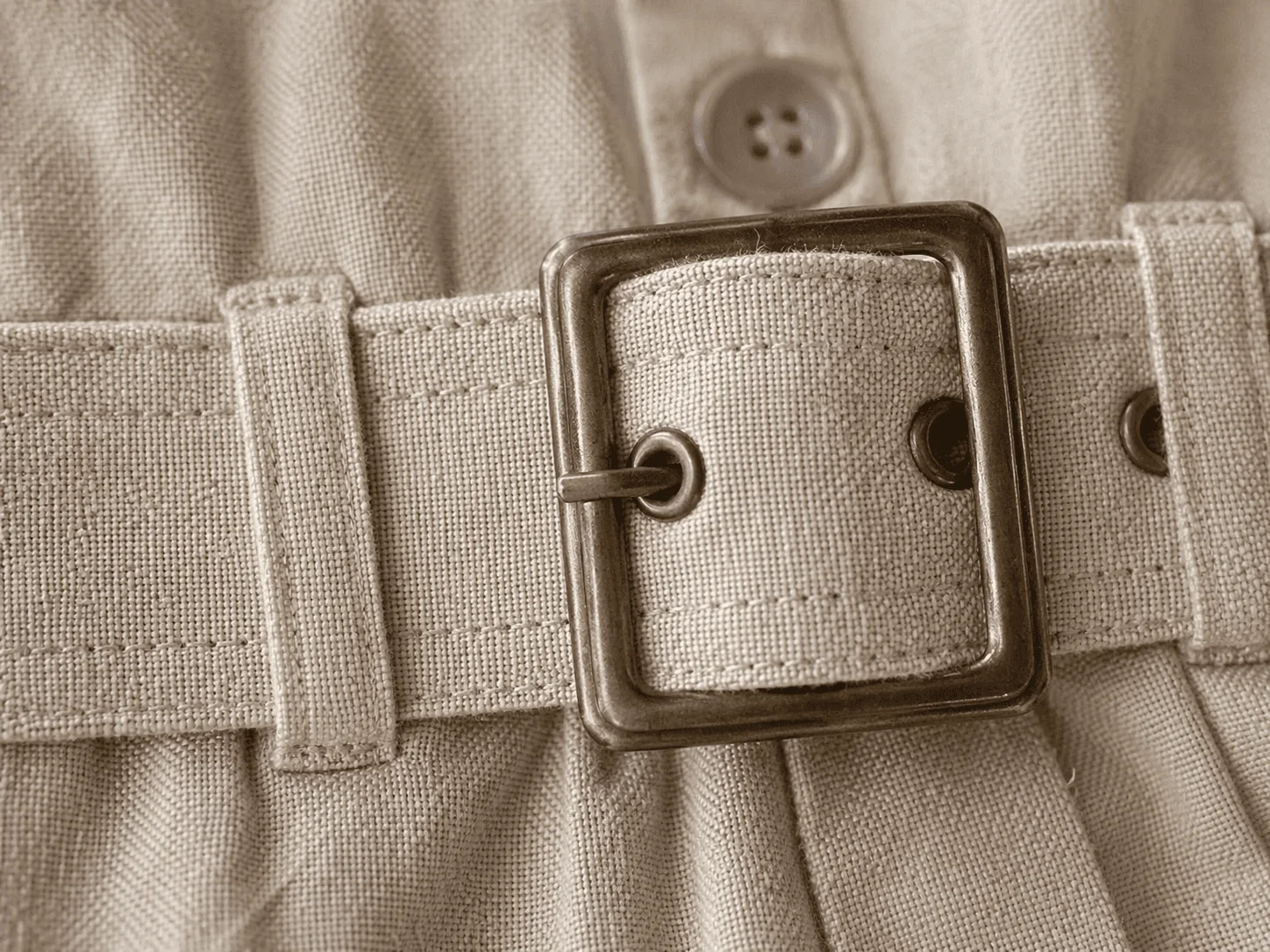 Macro do cós e cinto do Macacão Safari · fivela metálica em tom bronze envelhecido + ilhós metálicos ao lado · linho bege com pesponto duplo nas passadeiras · botão de metal acima · o aviamento que dá cara de roupa cara
