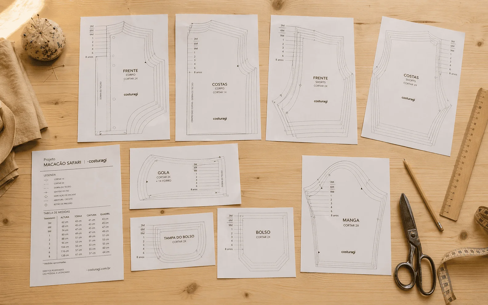Moldes do Macacão Safari impressos em papel sulfite sobre bancada de madeira — 8 peças identificáveis: Frente · Costas · Gola · Shortinho · Manga · Bolso · Tampa do bolso · capa 'Macação Safari' · régua e tesoura cegonha ao lado