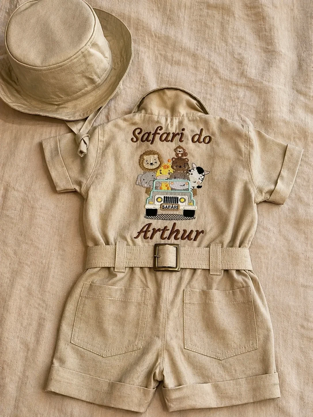 Flat-lay editorial do Macacão Safari visto pelas costas sobre fundo de linho cru · chapéu boonie no canto superior esquerdo · bordado central temático 'Safari do Arthur' em caligrafia marrom · Jipe branco com placa SAFARI transportando leão, macaco, animal escuro e zebra · cinto de linho com fivela quadrada bronze atravessando a cintura · dois bolsos traseiros com pesponto duplo · luz warm natural lateral · fotografia editorial Kinfolk
